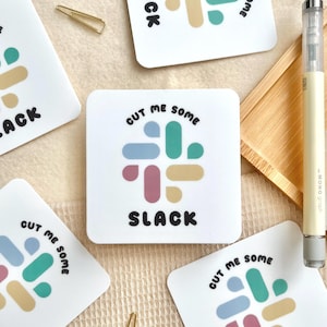 May include: White square coasters with a colorful abstract design and the words "CUT ME SOME SLACK". A gold paperclip and a pen are also visible.