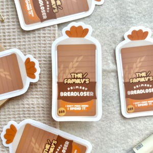 May include: Several stickers with a bread loaf design. The stickers feature the text "The Family's Primary Breadloser" and "I don't make bread, I spend it." The stickers have a brown and orange color scheme.
