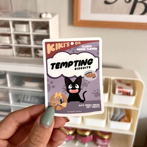 May include: A sticker featuring a cartoon black cat with a white mouth and white eyes. The sticker is a parody of a cat food package and reads "Kiki's Co. Fluffy Bread Flavor Tempting Biscuits". The sticker also features a cartoon bread and the text "Jiji's Fresh Baked Cat Treats".