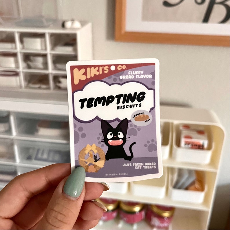 May include: A sticker featuring a cartoon black cat with a white mouth and white eyes. The sticker is a parody of a cat food package and reads "Kiki's Co. Fluffy Bread Flavor Tempting Biscuits". The sticker also features a cartoon bread and the text "Jiji's Fresh Baked Cat Treats".
