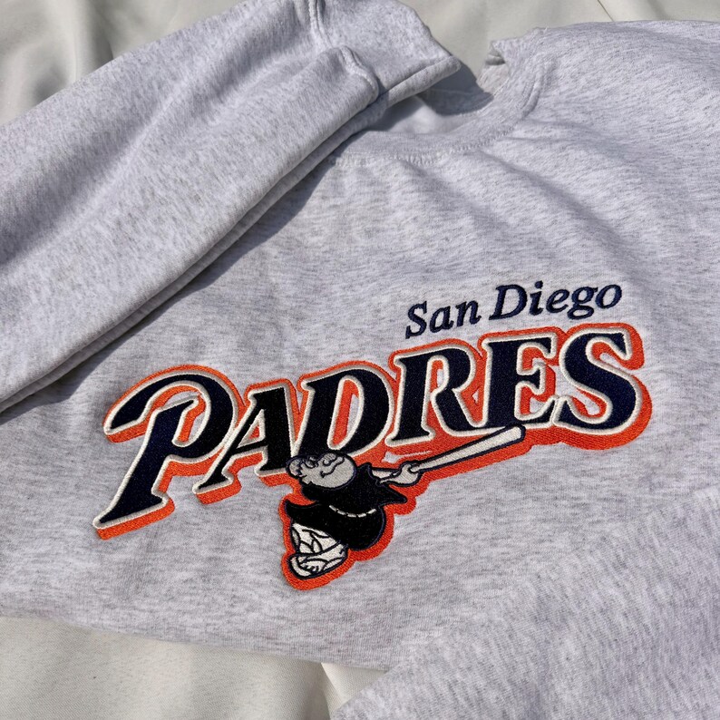 May include: A heather gray sweatshirt featuring the San Diego Padres logo. The logo includes the word "PADRES" in navy blue with an orange outline, and a cartoon character swinging a baseball bat. The words "San Diego" are above the logo.