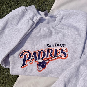 May include: A light gray sweatshirt featuring the San Diego Padres logo in navy blue, orange, and white. The logo includes the team name and a baseball player illustration. The sweatshirt is made of a soft material.