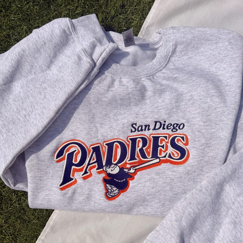 May include: A light gray sweatshirt featuring the San Diego Padres logo in navy blue, orange, and white. The logo includes the team name and a baseball player illustration. The sweatshirt is made of a soft material.