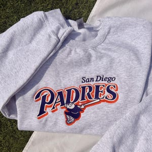 May include: A light gray sweatshirt featuring the San Diego Padres logo in navy blue, orange, and white. The logo includes the team name and a baseball player illustration. The sweatshirt is made of a soft material.
