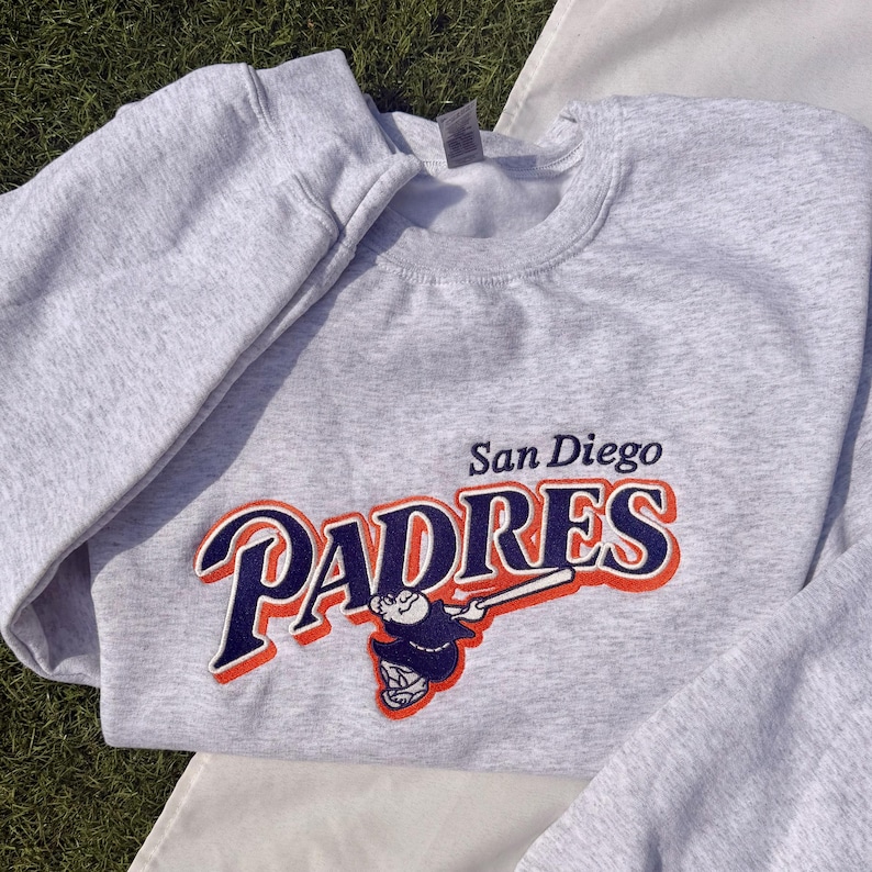 May include: A light gray sweatshirt featuring the San Diego Padres logo in navy blue, orange, and white. The logo includes the team name and a baseball player illustration. The sweatshirt is made of a soft material.