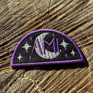 Puede incluir: Un parche bordado en morado y blanco con una luna creciente y la palabra "AMI" en letras moradas. El parche está rodeado de pequeñas estrellas blancas sobre un fondo negro.