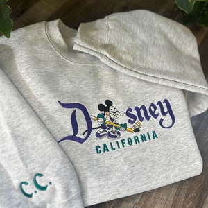 Op de afbeelding: Grijs sweatshirt met een geborduurd ontwerp in paars en groen van Mickey Mouse die hockey speelt. De tekst "Disney California" is onder de afbeelding geborduurd.