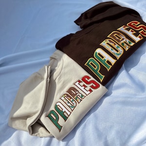 May include: Two hooded sweatshirts with the word "PADRES" embroidered on the sleeve in green, red, and white with a Mexican flag design. The sweatshirts are brown and beige.