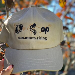 May include: A beige baseball cap featuring black embroidered zodiac symbols for Pisces, Aquarius, and Aries, along with the text "sun.moon.rising" stitched in black thread across the front panel above the curved brim.