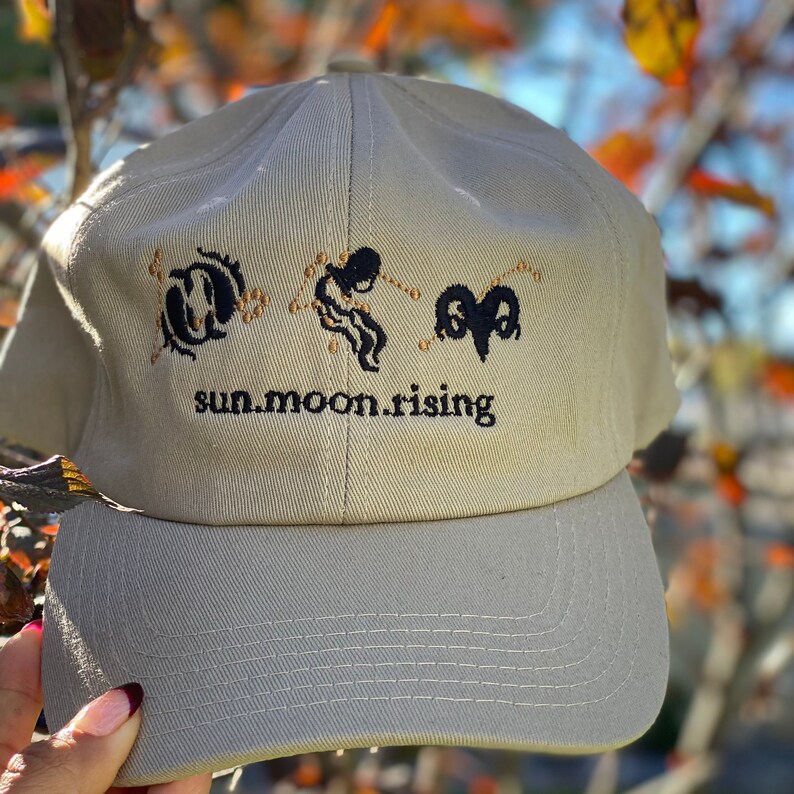 May include: A beige baseball cap featuring black embroidered zodiac symbols for Pisces, Aquarius, and Aries, along with the text "sun.moon.rising" stitched in black thread across the front panel above the curved brim.