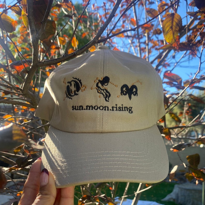 May include: A beige baseball cap featuring black embroidered zodiac symbols and the text "sun.moon.rising" in a lowercase serif font. The design includes three distinct astrological icons arranged horizontally above the text on the front panel.