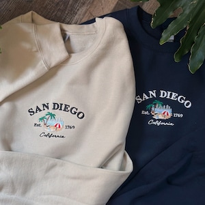 Puede incluir: Dos sudaderas con diseños bordados de San Diego. La sudadera de la izquierda es beige con una palmera y una escena de playa. La sudadera de la derecha es azul marino con un diseño similar.
