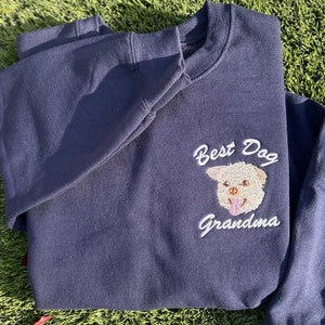 May include: A navy blue sweatshirt with white embroidery that reads "Best Dog Grandma" and features a white dog with a pink tongue.