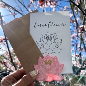 Op de afbeelding: Een witte kaart met de tekst "lotus flower" en een zwarte lijntekening van een lotusbloem. Op de voorgrond ligt een roze geborduurde lotusbloem patch.