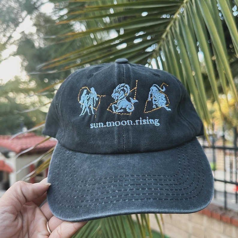 May include: A charcoal gray baseball cap featuring light blue embroidered zodiac symbols and constellations. Below the embroidery, the text "sun.moon.rising" is stitched in light blue thread. The cap has a curved brim and a structured crown.