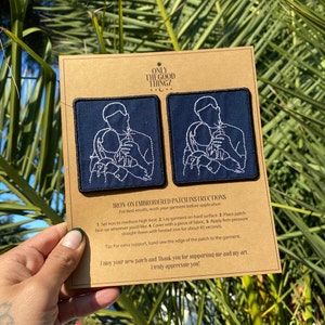 Op de afbeelding: Twee geborduurde strijkpatches op een bruine kaart. De patches zijn donkerblauw met witte borduursels van een stel dat elkaars hand vasthoudt. De kaart bevat instructies voor het aanbrengen van de patches. De tekst op de kaart luidt "Only the Good Thingz" en "Instructies voor geborduurde strijkpatches".