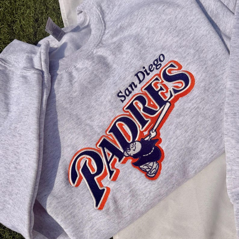 May include: A light gray sweatshirt featuring the San Diego Padres logo in navy blue, orange, and white. The logo includes the word "PADRES" and a baseball player swinging a bat. The sweatshirt is made of a soft material.