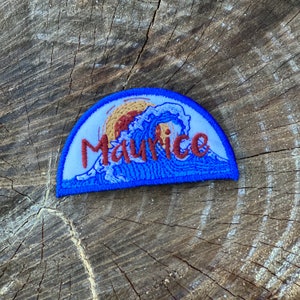 Puede incluir: Un parche bordado azul y blanco con una ola y la palabra "Maurice" en letras marrones.