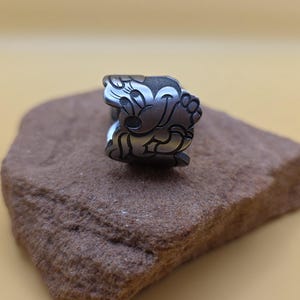 Op de afbeelding: Een zilverkleurige ring met een cartoonkarakterontwerp. De ring toont een lachend karakter met grote oren en een vrolijke uitdrukking. De ring is geplaatst op een bruine, getextureerde steen tegen een gele achtergrond.