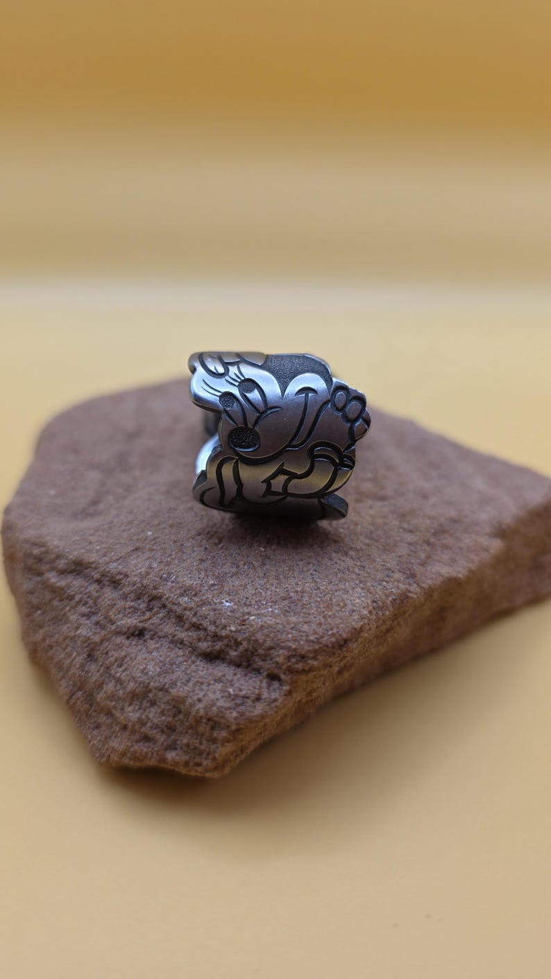 Op de afbeelding: Een zilverkleurige ring met een cartoonkarakterontwerp. De ring toont een lachend karakter met grote oren en een vrolijke uitdrukking. De ring is geplaatst op een bruine, getextureerde steen tegen een gele achtergrond.