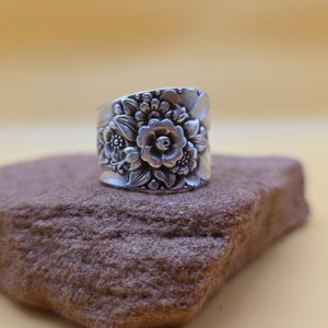 May include: A wide silver-toned ring crafted from a repurposed spoon, featuring an intricate embossed floral pattern with detailed petals and leaves covering the entire outer surface of the band, showcasing a polished metallic finish.