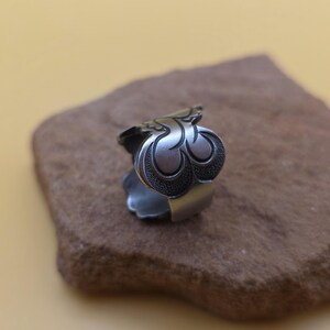 Op de afbeelding: Een zilverkleurige ring met een sierlijk, gestileerd bloemmotief. De ring heeft een gedetailleerd, drielobbig motief op de voorkant, met een getextureerd, donker patroon in de lobben. De ring ligt op een bruin stenen oppervlak.
