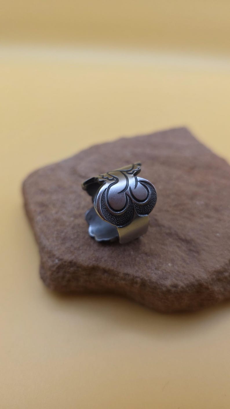 Op de afbeelding: Een zilverkleurige ring met een sierlijk, gestileerd bloemmotief. De ring heeft een gedetailleerd, drielobbig motief op de voorkant, met een getextureerd, donker patroon in de lobben. De ring ligt op een bruin stenen oppervlak.