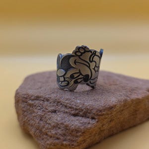 Op de afbeelding: Een zilverkleurige ring met een gedetailleerd cartoonkarakterontwerp. De ring is geplaatst op een kleine, getextureerde, bruine steen tegen een effen gele achtergrond. Het ontwerp van de ring toont een gezicht met grote oren en een lachende uitdrukking.