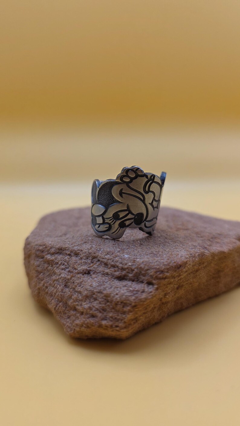 Op de afbeelding: Een zilverkleurige ring met een gedetailleerd cartoonkarakterontwerp. De ring is geplaatst op een kleine, getextureerde, bruine steen tegen een effen gele achtergrond. Het ontwerp van de ring toont een gezicht met grote oren en een lachende uitdrukking.