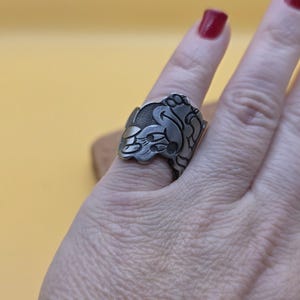 Op de afbeelding: Een zilverkleurige ring met een sierlijk bloemen- en bladerontwerp. De ring wordt gedragen aan een vinger met rode nagellak. Het ontwerp is gedetailleerd en lijkt handgemaakt.