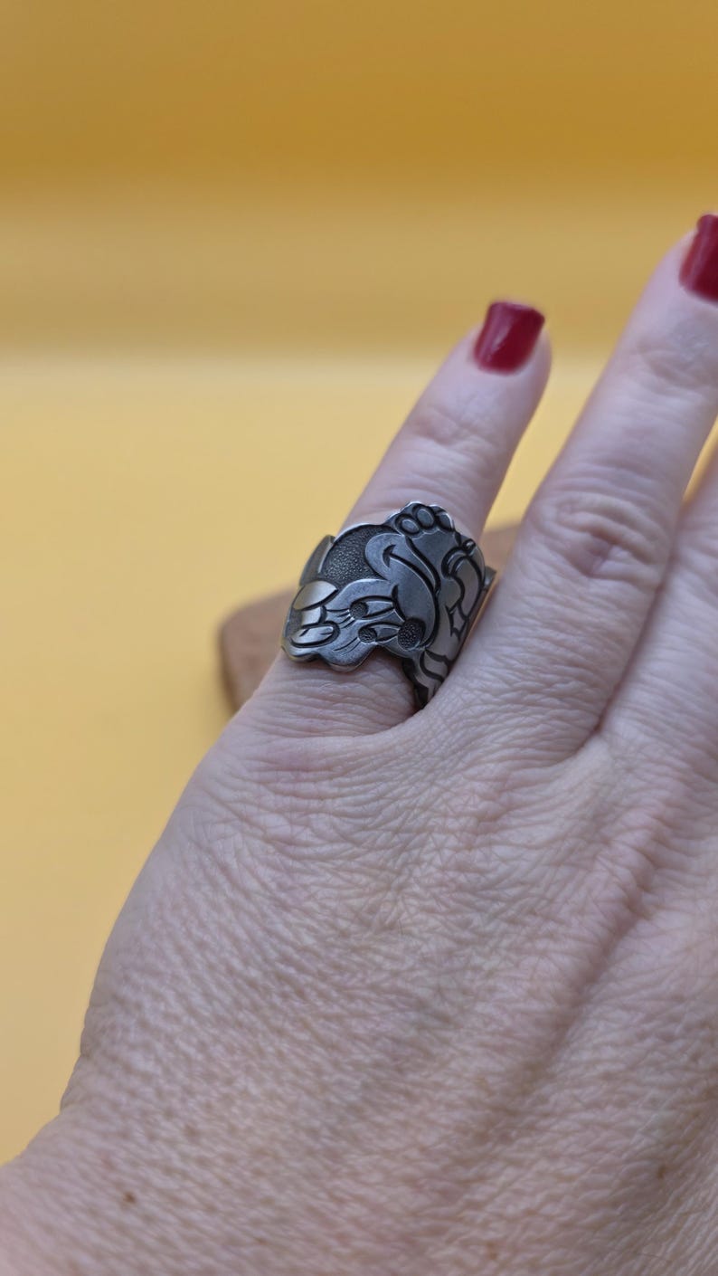 Op de afbeelding: Een zilverkleurige ring met een sierlijk bloemen- en bladerontwerp. De ring wordt gedragen aan een vinger met rode nagellak. Het ontwerp is gedetailleerd en lijkt handgemaakt.