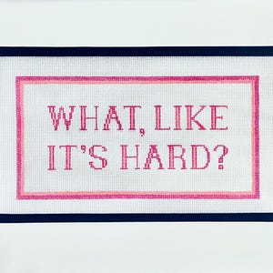 May include: A needlepoint canvas with the phrase "WHAT, LIKE IT'S HARD?" stitched in pink. The text is centered on a white background, framed by a pink and navy blue border.
