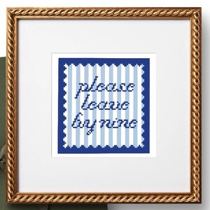 May include: A framed cross-stitch artwork with the words "please leave by nine" in blue thread. The background features vertical blue and white stripes. The artwork is set in a gold-colored, ornate frame.