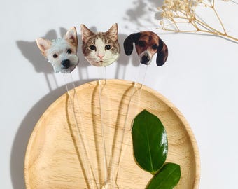 Könnte beinhalten: Drei klare Cocktailrührer mit Tiergesichtern: ein weißer Hund, eine Katze und ein brauner Hund. Die Rührer sind auf einem Holztablett mit grünen Blättern angeordnet. Der Hintergrund ist weiß, mit einem Zweig getrockneter Blumen.