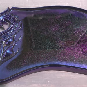 May include: A decorative tray with a dragon head design. The tray is a deep purple colour with a glittery, iridescent finish. The dragon head is on the left side, with intricate details. The tray has a curved shape.