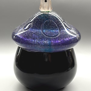 Könnte beinhalten: Ein schwarzer, pilzförmiger Behälter mit einem glitzernden, lila und blauen Deckel. Eine kleine, durchsichtige Geisterfigur sitzt auf dem Deckel. Der Behälter hat eine glänzende Oberfläche, und das Gesamtdesign ist skurril und dekorativ.
