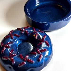 May include: A navy blue donut-shaped container with dark red sprinkles next to a matching navy blue bowl. The donut container has a glossy finish, and the sprinkles are evenly spaced across the top.