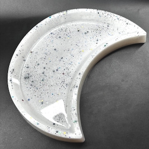 May include: A white, crescent moon-shaped tray with embedded glitter. The tray has a raised edge and a glossy finish. The glitter is a mix of silver, black, and iridescent colours, creating a sparkling effect. The tray is designed for decorative use, perhaps for jewellery.