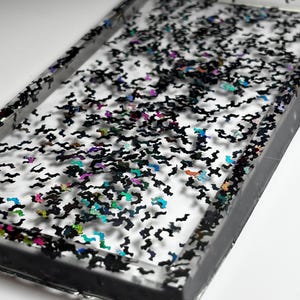 May include: A rectangular tray made of clear resin featuring a dense scattering of small, black, bat-shaped glitter pieces. The glitter includes iridescent accents that reflect various colors, and the tray has a solid black border along the edges.