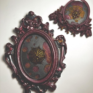 May include: Two oval-shaped decorative frames in a dark metallic burgundy finish with ornate, swirling edges. Each frame contains a black background with shimmering copper-colored spots and a central gold-colored relief of a spider web.