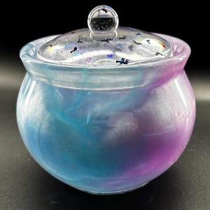 Puede incluir: Un pequeño tarro redondo con tapa transparente y asa pequeña. El tarro es un degradado de azul y morado, con efecto veteado. La tapa es transparente con pequeñas formas oscuras incrustadas.