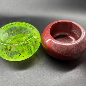 May include: Close-up of three resin bowls in varying colours. The first is a transparent green with glitter, the second is a solid maroon, and the third is a marbled pink and white. The bowls are round with a smooth, glossy finish.