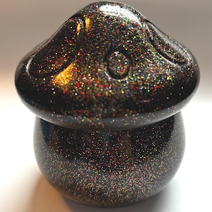 May include: A black mushroom-shaped container with a lid, covered in iridescent glitter. The container has a rounded base and a mushroom cap-shaped lid with a small circular indentation. The glitter reflects various colors.