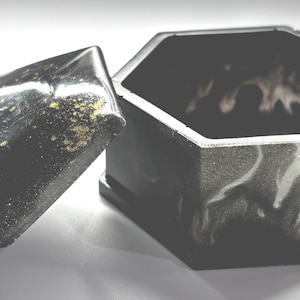 Könnte beinhalten: Eine sechseckige schwarze Box mit leicht geöffnetem Deckel. Die Box und der Deckel haben ein marmoriertes Aussehen mit Wirbeln aus Grau und Goldflocken. Das Innere der Box ist dunkel, und der Deckel ist nach links abgewinkelt.