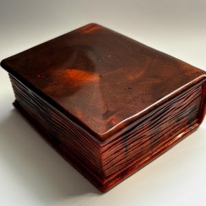May include: A decorative book-shaped box with a glossy, reddish-brown finish. The box is designed to resemble a closed book, with detailed edges mimicking pages. The top surface has a marbled effect, adding visual interest. The box is a unique storage item.