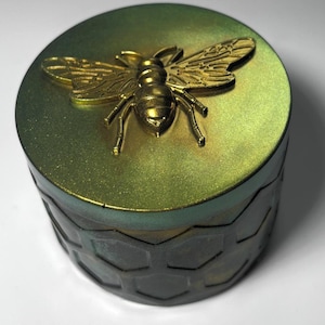 Pode incluir: Um recipiente redondo e decorativo com uma abelha dourada na tampa. A tampa e as laterais têm um efeito degradê verde e dourado. As laterais do recipiente apresentam um padrão de favo de mel.