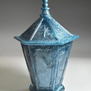 Pode incluir: Um pote hexagonal com tampa em acabamento azul marmorizado brilhante. O pote apresenta um padrão em relevo ornamentado em cada painel e um remate pontiagudo na tampa. Um emblema em forma de estrela está gravado na frente da tampa.