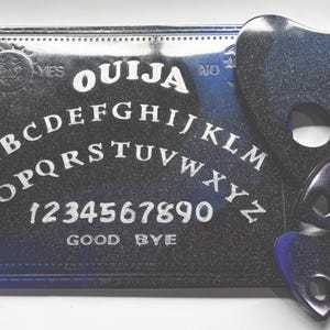 May include: A black Ouija board with white lettering and three planchette pieces. The board features the alphabet, numbers, and the words "YES", "NO", and "GOOD BYE". The planchette pieces are heart-shaped.