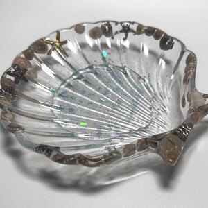 Puede incluir: Un plato de resina transparente con forma de concha que presenta pequeñas conchas marinas y estrellas de mar metálicas incrustadas a lo largo del borde festoneado. El interior está infundido con finos brillos iridiscentes que captan la luz, creando un efecto brillante.