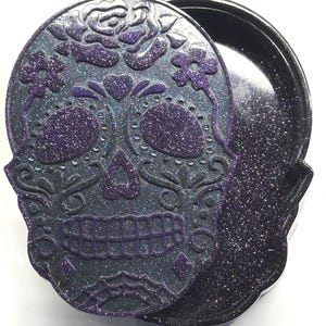 Puede incluir: Una caja decorativa con forma de calavera con tapa. La tapa presenta un diseño detallado de calavera de azúcar con detalles de purpurina morada. La caja es negra con purpurina, y la tapa está ligeramente entreabierta.